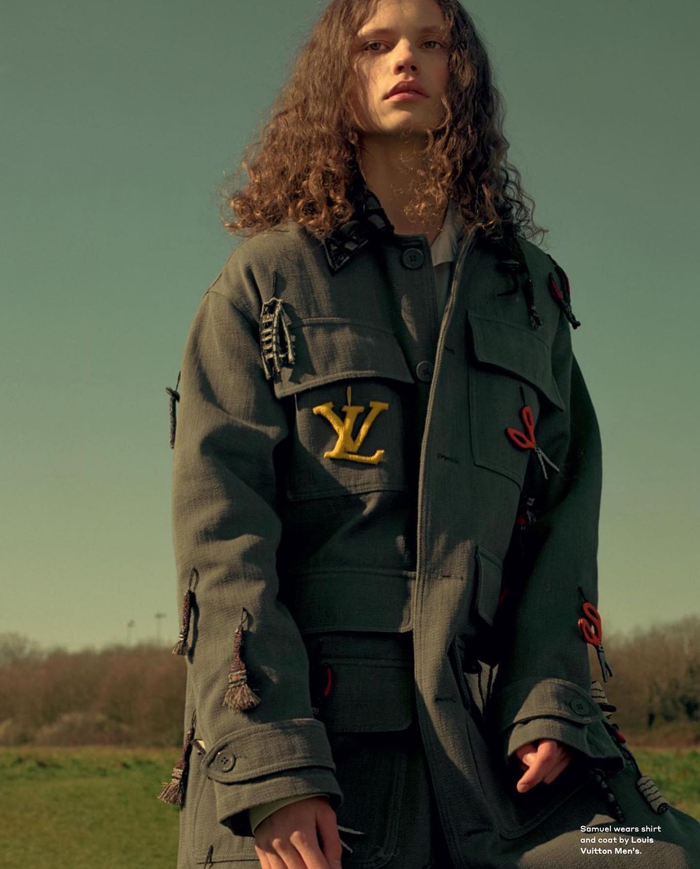 A young adult male fashion model with long brown hair poses in an outdoor field wearing an olive green Louis Vuitton jacket.