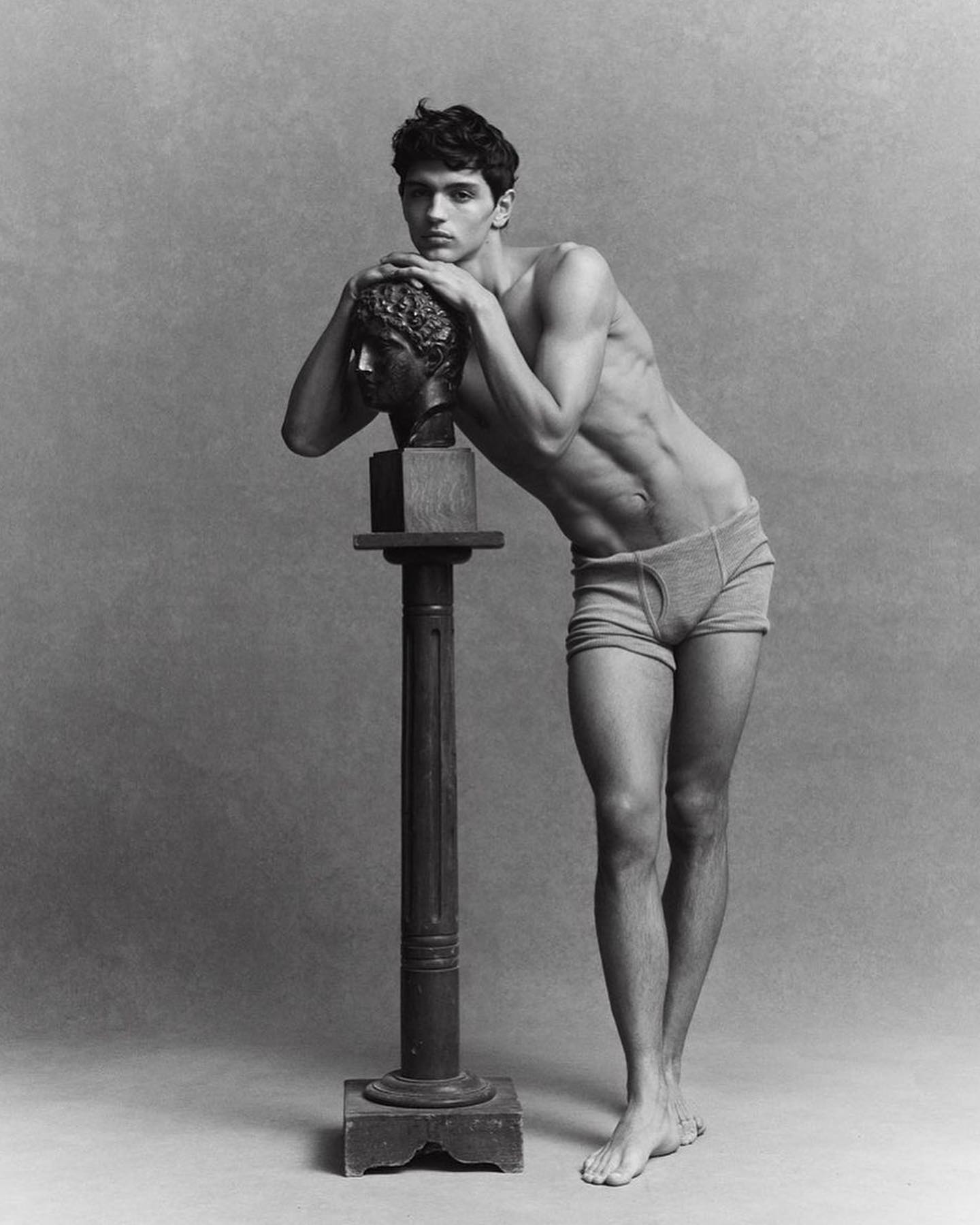 Black and white studio shot of a young man with a slim, muscular build leaning on a classical sculpture bust on a column.