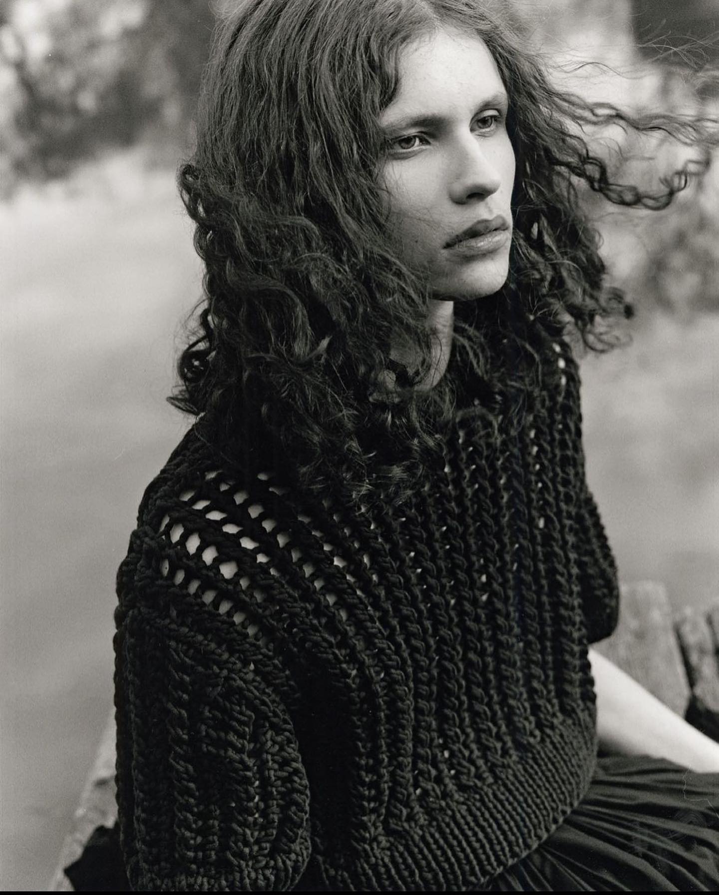 Monochrome portrait of a woman with curly hair wearing a knit top outdoors near a lake, with shallow depth of field.