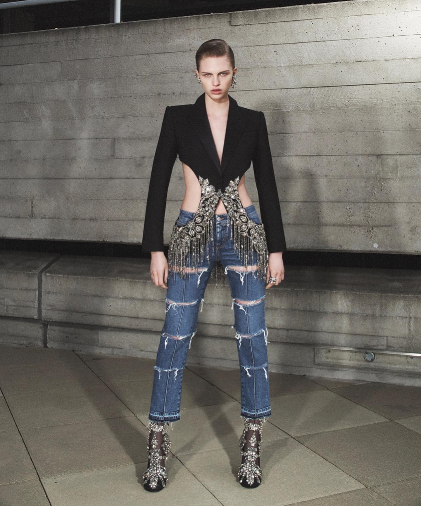 A slender, fair-skinned model poses in ripped jeans, a black blazer with cutouts, and embellished heels against a concrete wall.
