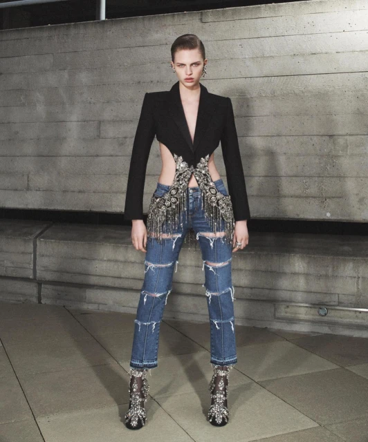 A slender, fair-skinned model poses in ripped jeans, a black blazer with cutouts, and embellished heels against a concrete wall.