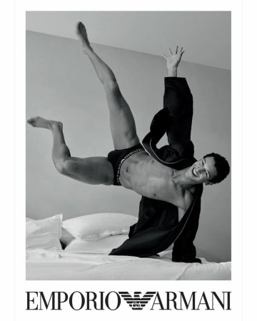 A smiling male model in underwear falling backward onto a bed in black and white.