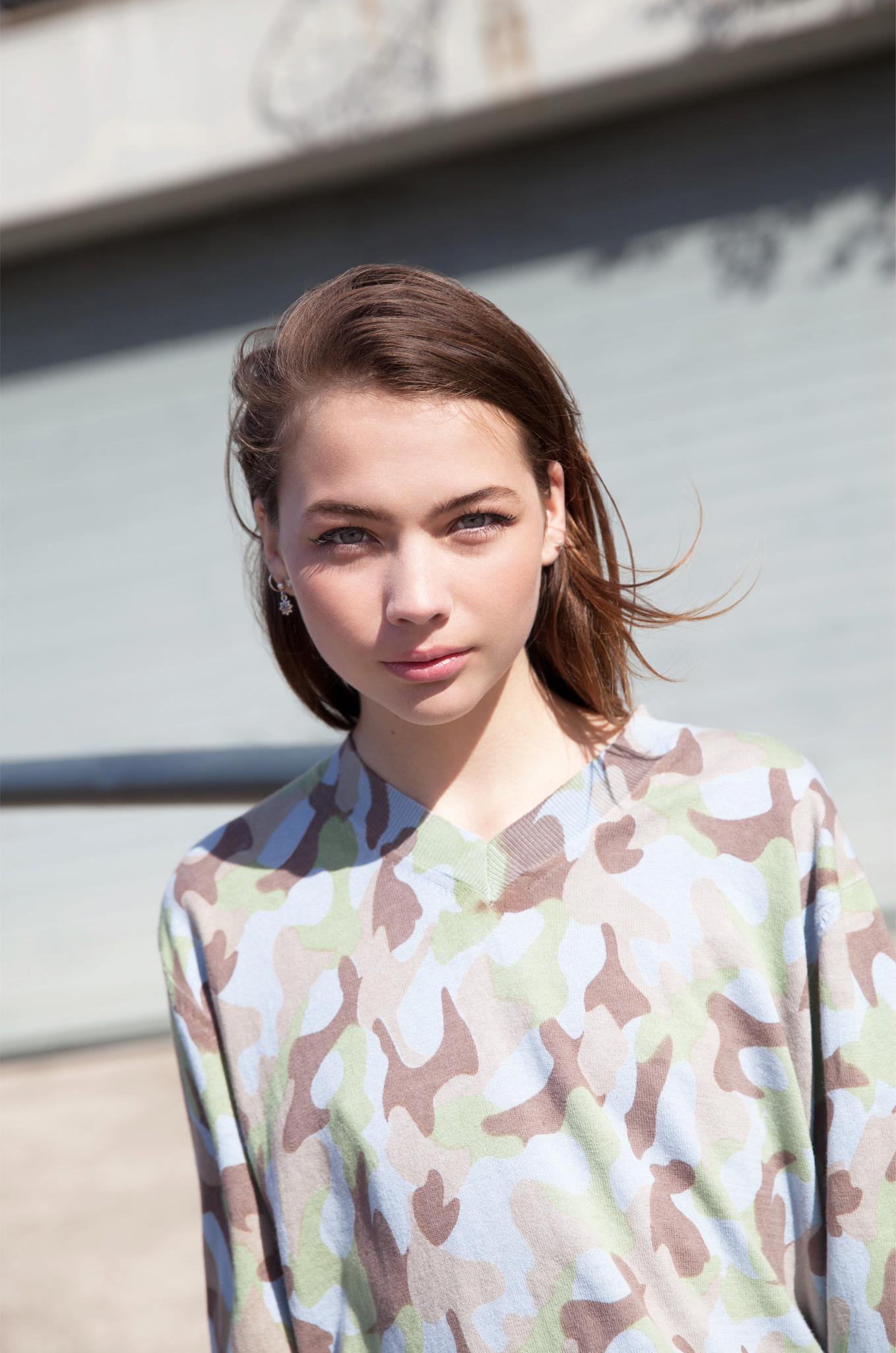 Portrait of a young woman with brown hair and blue eyes wearing a camouflage patterned shirt outdoors against a blurred building background.