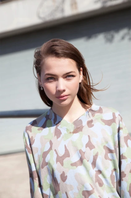 Portrait of a young woman with brown hair and blue eyes wearing a camouflage patterned shirt outdoors against a blurred building background.