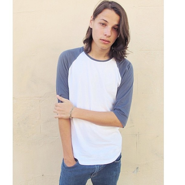 Portrait of a young adult man with long brown hair, wearing a two-tone t-shirt and jeans, posing against a beige wall.