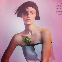 Model with short dark bob and blunt bangs wearing a strapless top featuring tucked flowers against a pink and white gradient backdrop.