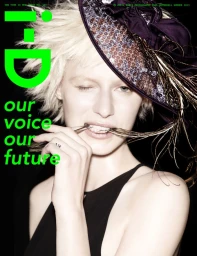 Close-up portrait of a blonde model with spiked hair wearing a dark fascinator hat and black top against a black background.