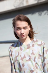 Close-up portrait of a young woman with brown hair wearing a blue and green camouflage pattern top outdoors.