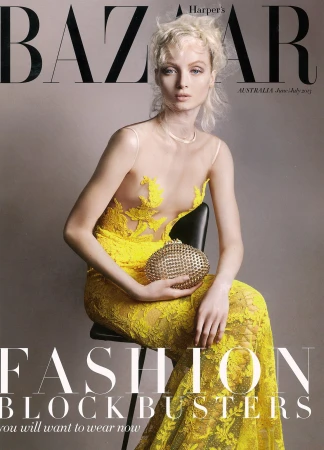 Model with blonde hair seated on a stool wearing a yellow lace gown holding an embellished gold clutch handbag against grey background
