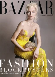 Model with blonde hair seated on a stool wearing a yellow lace gown holding an embellished gold clutch handbag against grey background