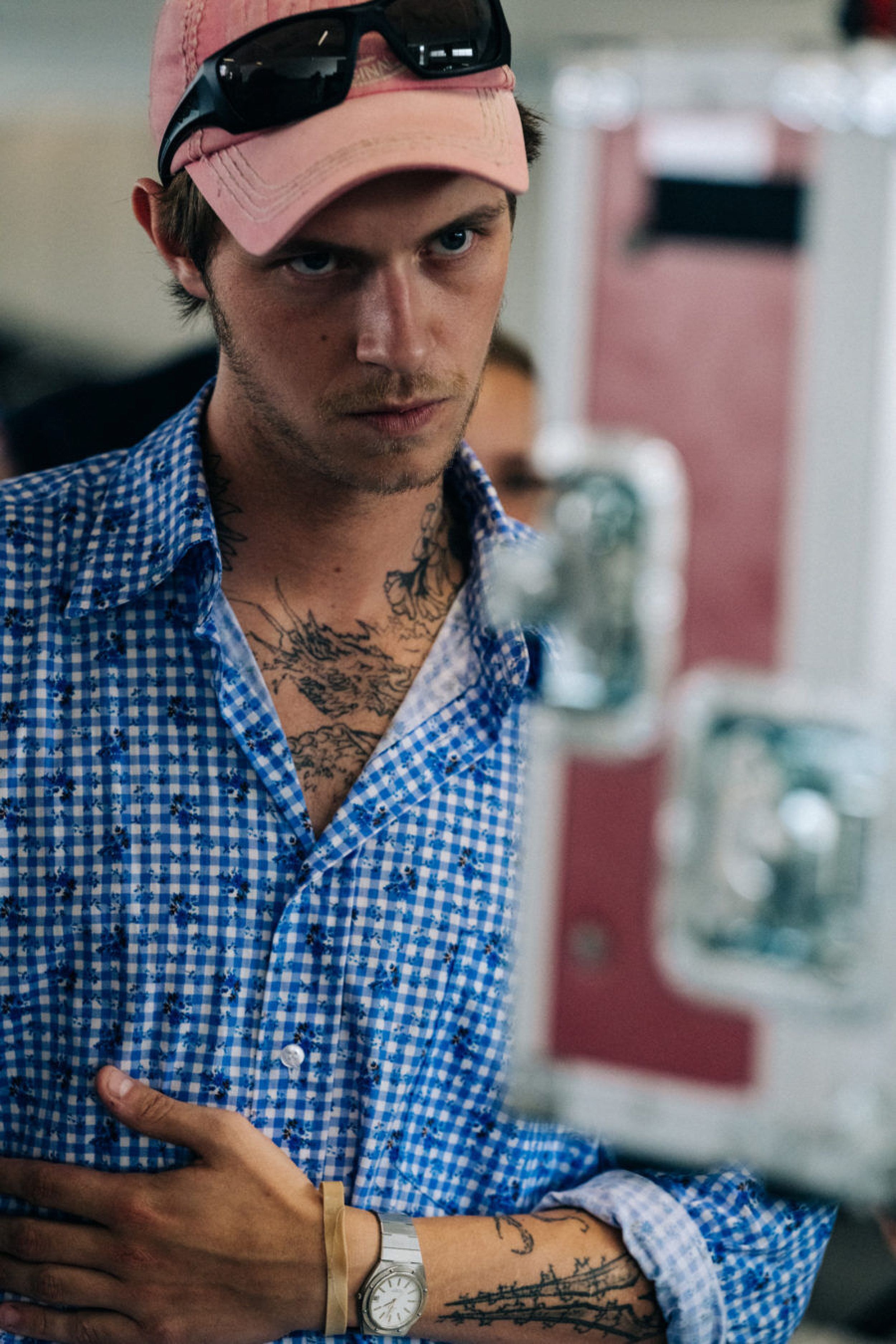 Close-up shot of a fair-skinned adult male with tattoos on his neck and arm, wearing a blue patterned shirt and a pink baseball cap.