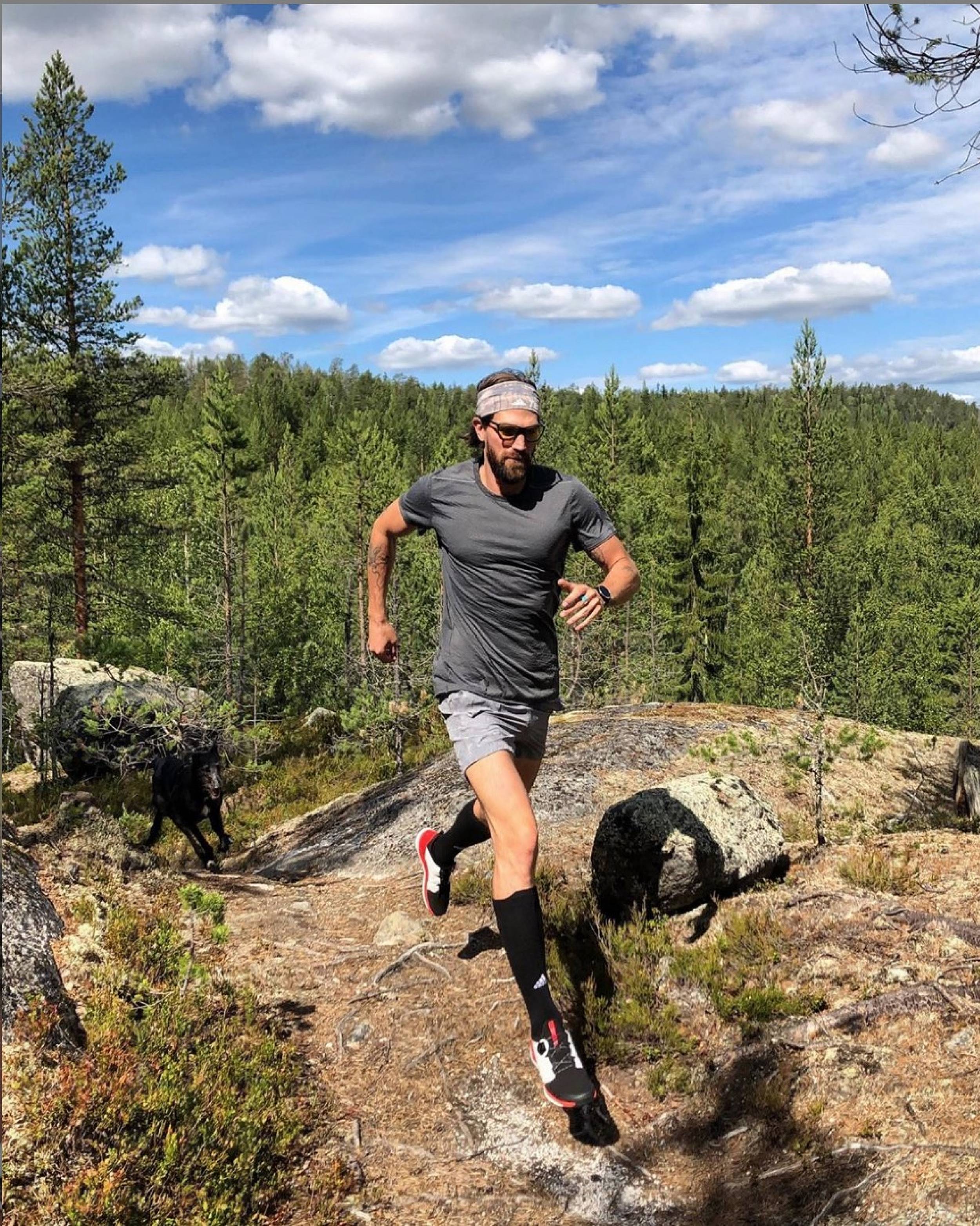 Athletic man running on a trail outdoors, wearing athletic wear, with a dog in the background, trees and blue sky overhead.
