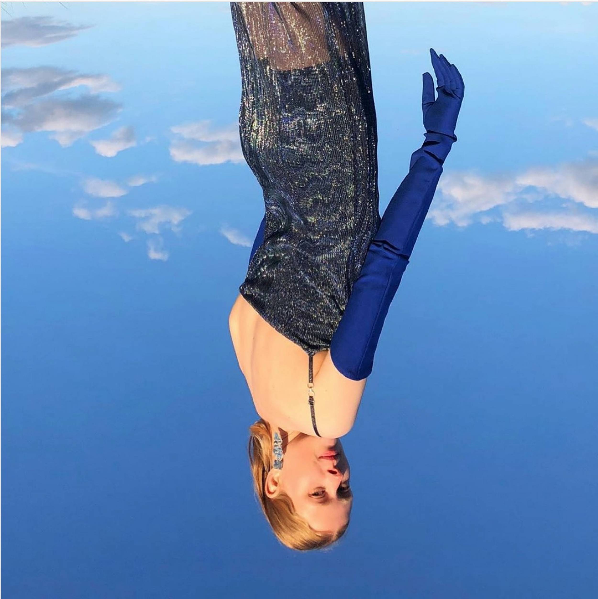 A woman in an iridescent dress and blue gloves poses against a blue sky with light clouds, in an eye-level outdoor shot.