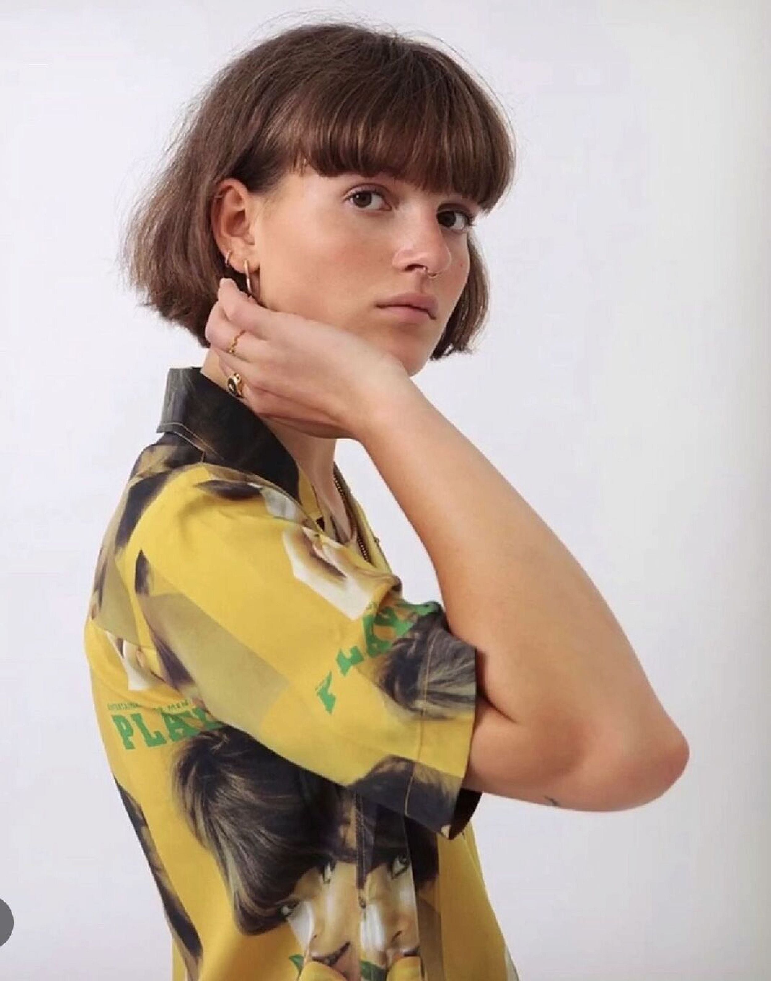 Portrait of a young woman with short brunette hair and yellow patterned shirt posing against a white backdrop in a photo studio.
