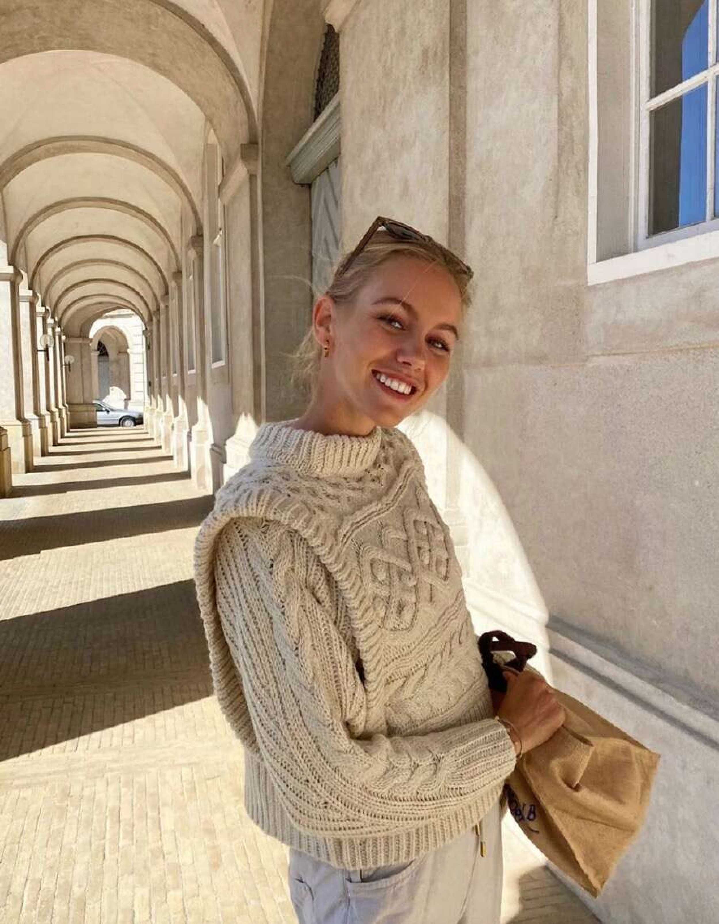 Smiling young woman with sunglasses poses in front of an arched building wearing a cream-colored cable knit sweater and holding a bag.