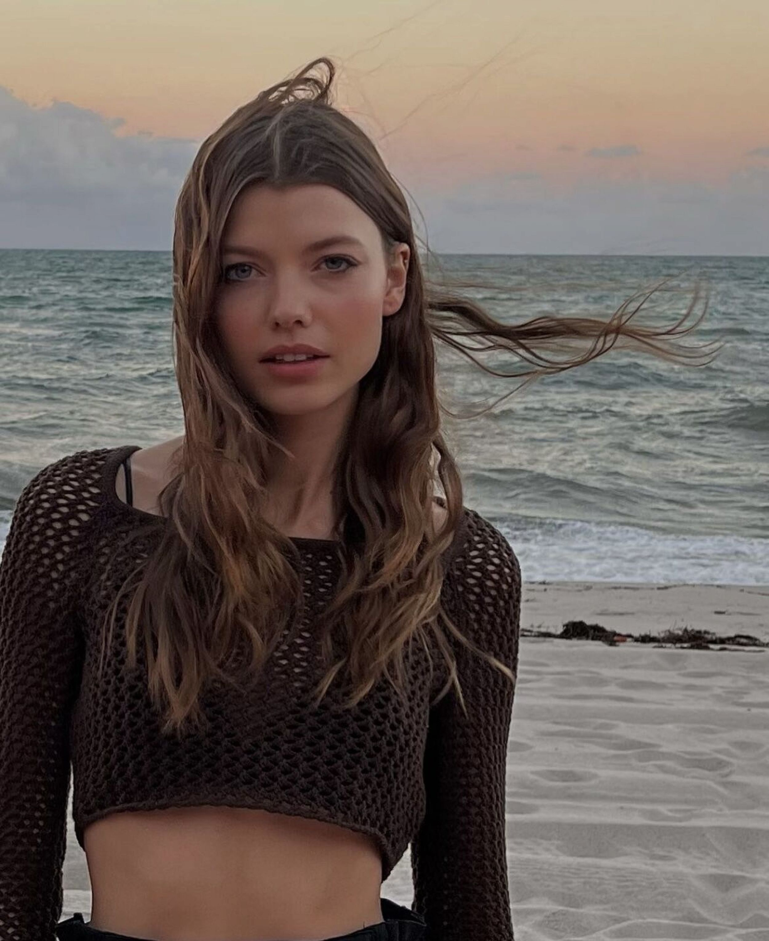 Woman in a brown crochet top stands on a sandy beach with the ocean and a pastel sky in the background during the daytime.