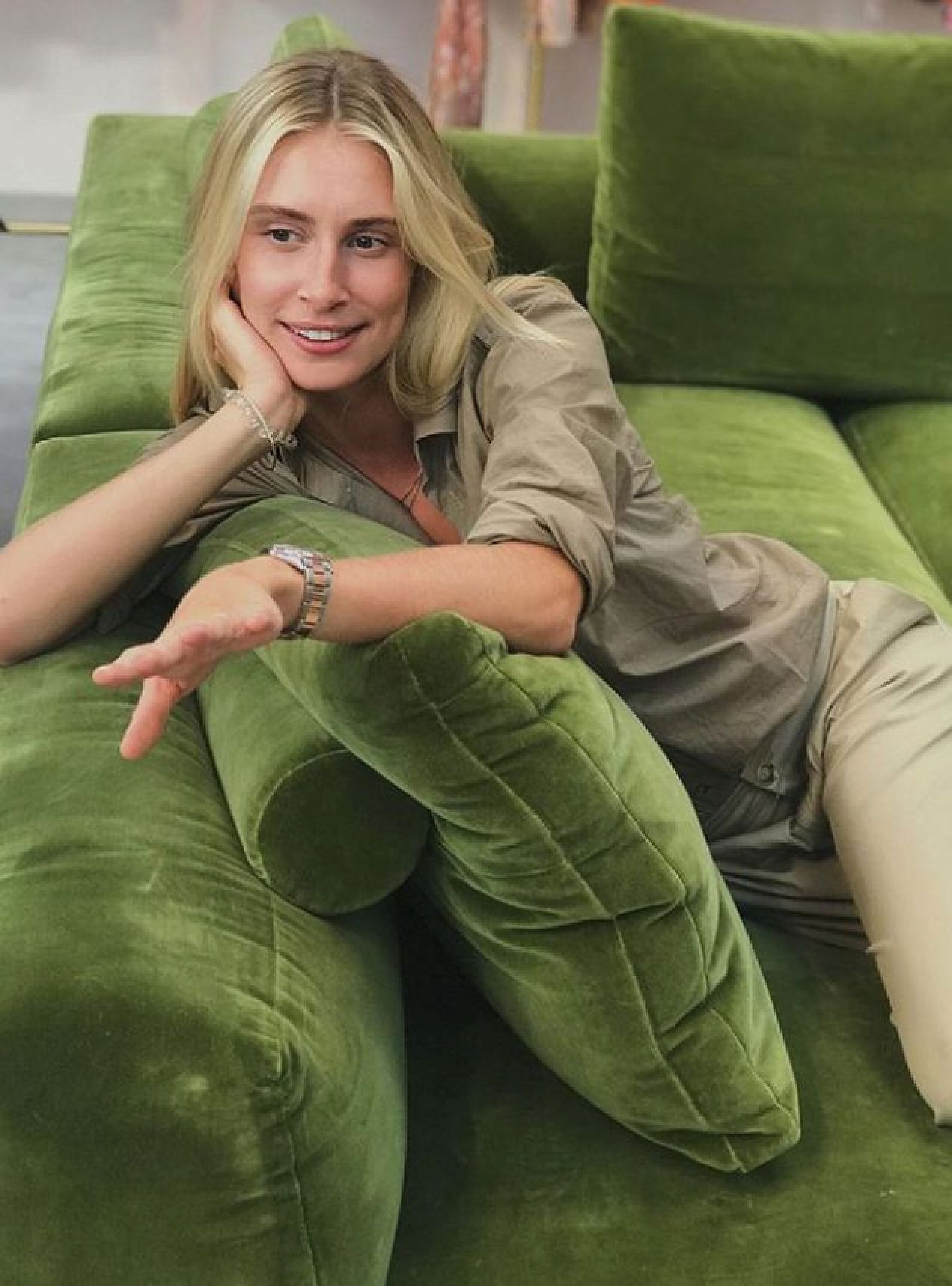 Adult woman with blonde hair lounges casually on a green velvet couch in an indoor setting, looking off to the side and smiling.