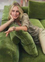 Blonde woman wearing khaki shirt and trousers reclining on a green velvet sofa, resting chin on hand