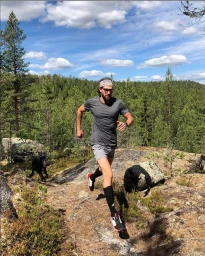 Man running on a forest trail with a dog, wearing grey athletic clothing, long socks, and a headband