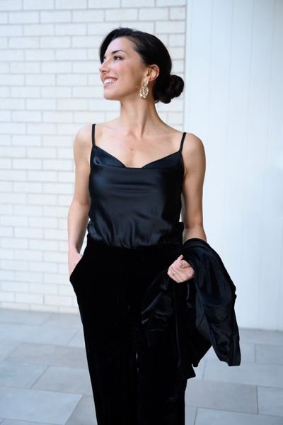 Elegant woman in a black camisole and velvet pants poses against a white brick wall, holding a black jacket and smiling.