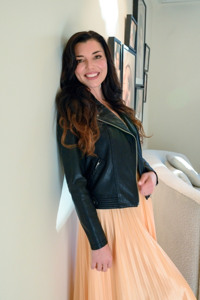 Portrait of a smiling brunette woman in a black leather jacket and pleated peach skirt posing against an indoor wall near framed art.