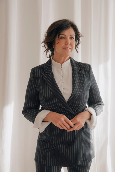 Adult woman in a pinstripe suit stands indoors near a window with a white curtain, hands clasped, in a medium shot with soft, diffused lighting.