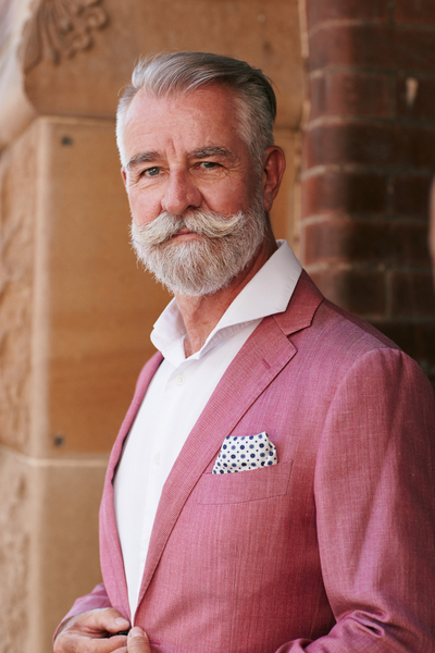 Adult man with grey hair and handlebar mustache poses wearing a pink linen blazer and white shirt in front of a stone wall.