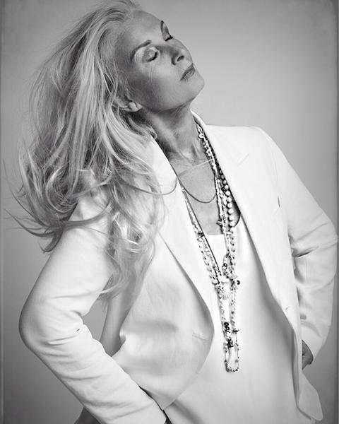 Elegant older woman with long silver hair and white blazer poses with hands on hips against a neutral background in a black-and-white studio portrait.