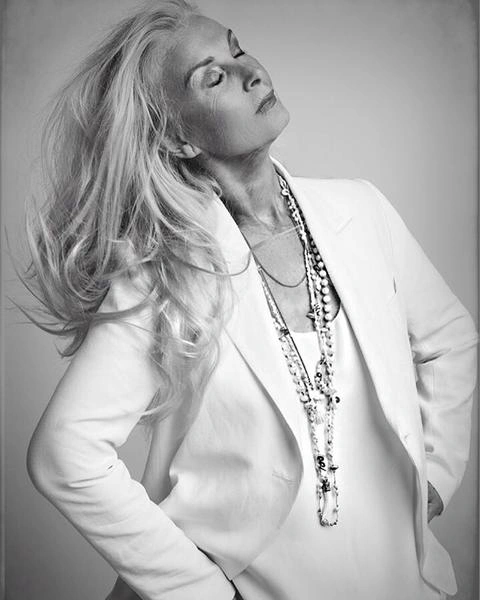 Elderly woman with long white hair wearing a white blazer and layered necklaces against a plain studio background.