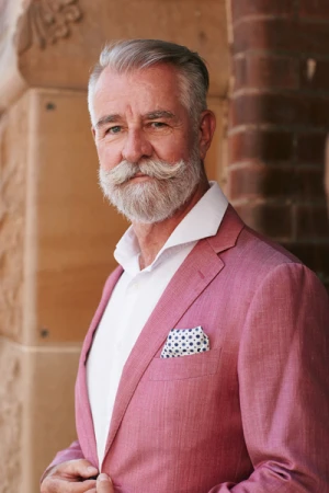 Elderly man with white beard wearing a pink blazer and white shirt standing against a brick building exterior.