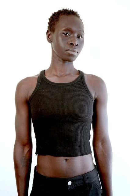 Portrait of a dark-skinned adult female in a black ribbed crop top and dark pants against a bright white studio background.