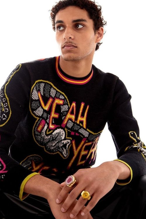 A stylish young adult poses in a studio, wearing a black sweater featuring a snake and the word 'Yeah,' complemented by gold rings.