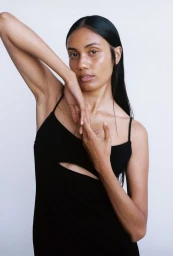 A woman with long dark hair wearing a black spaghetti strap dress posing against a plain white background.