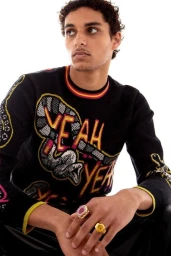 A young man wearing a black knit sweater with graphic text and snake motifs, posing against a solid white background.