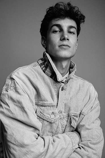 Monochrome portrait of a young adult man wearing a denim jacket with arms crossed, set against a neutral gray backdrop in a studio.