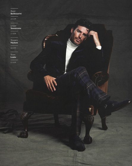 A distinguished man is seated in a wingback chair wearing a black blazer, white turtleneck, and patterned trousers in a fashion setting.