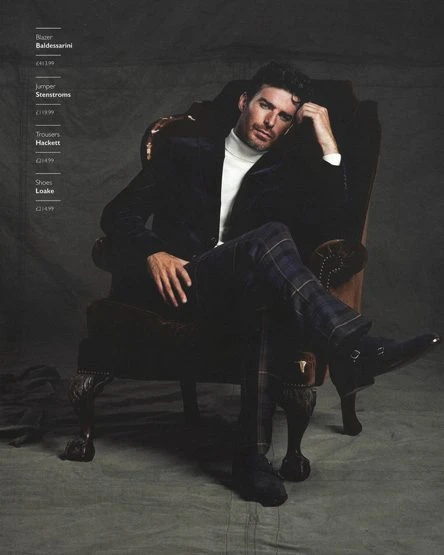 A distinguished man is seated in a wingback chair wearing a black blazer, white turtleneck, and patterned trousers in a fashion setting.