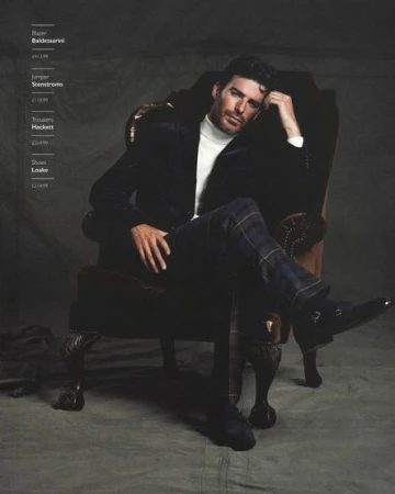 A distinguished man is seated in a wingback chair wearing a black blazer, white turtleneck, and patterned trousers in a fashion setting.