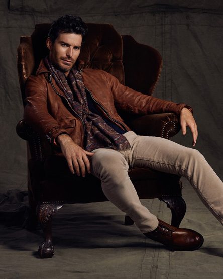 Adult male in brown leather jacket and scarf sitting in an armchair in a studio with neutral backdrop and warm-toned lighting.