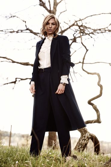 A stylish woman with blonde hair poses in a navy coat and pants outdoors against a backdrop of dry grass and bare tree branches.