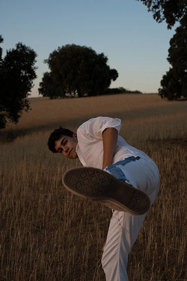 A young adult poses outdoors in a field at dusk, wearing casual white clothing and striking a kicking pose with his leg extended.