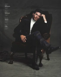 A man in a velvet blazer and plaid trousers seated in an armchair against a textured dark backdrop.