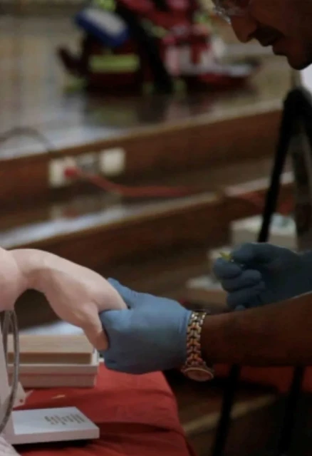 Healthcare provider wearing blue gloves holds a patient's hand inside of a clinic. Visible medical equipment and supplies in background.