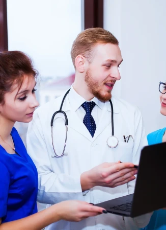 Three healthcare workers in blue scrubs and white coats look at a laptop indoors, discussing a patient's case.