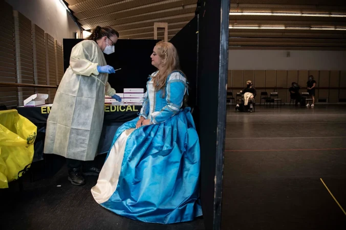 Medical professional administering COVID-19 test to woman in period costume in indoor testing location with other individuals in the background.
