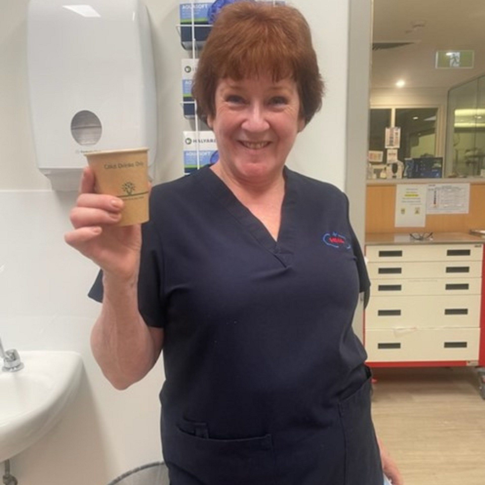 A smiling woman in a blue medical uniform holds a disposable coffee cup in a bright indoor healthcare setting, looking directly at the camera.