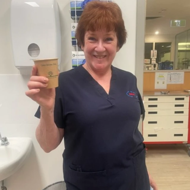 A smiling woman in a blue medical uniform holds a disposable coffee cup in a bright indoor healthcare setting, looking directly at the camera.