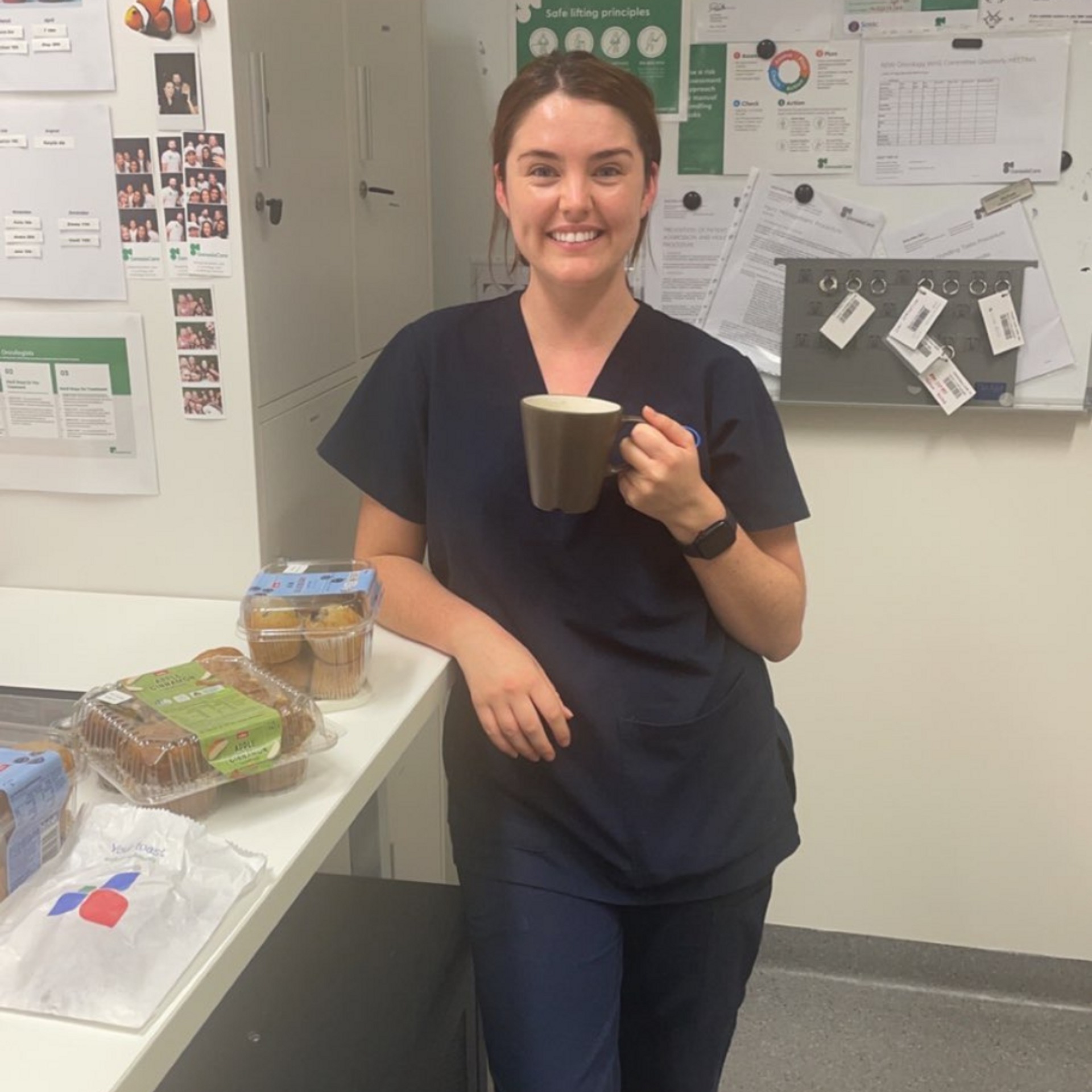 Smiling woman in scrubs enjoys a coffee break with muffins in what looks like a healthcare or office break room setting.