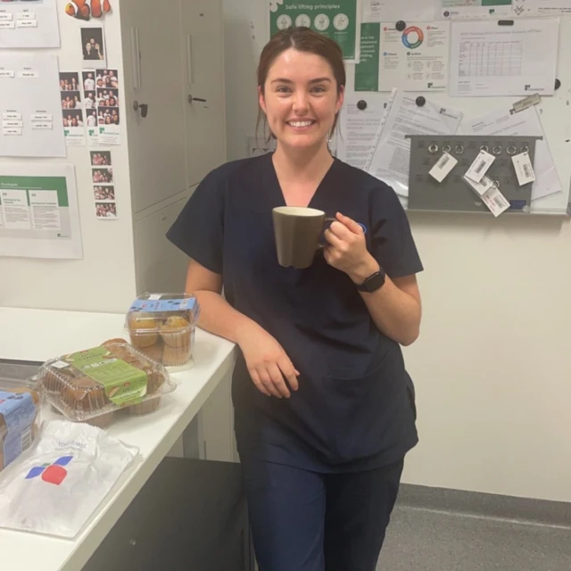 Smiling woman in scrubs enjoys a coffee break with muffins in what looks like a healthcare or office break room setting.