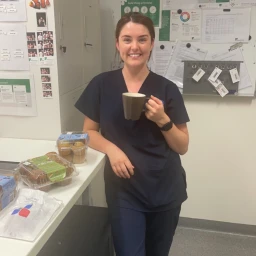 Smiling female nurse in navy scrubs holding a brown mug inside an office breakroom in Sydney